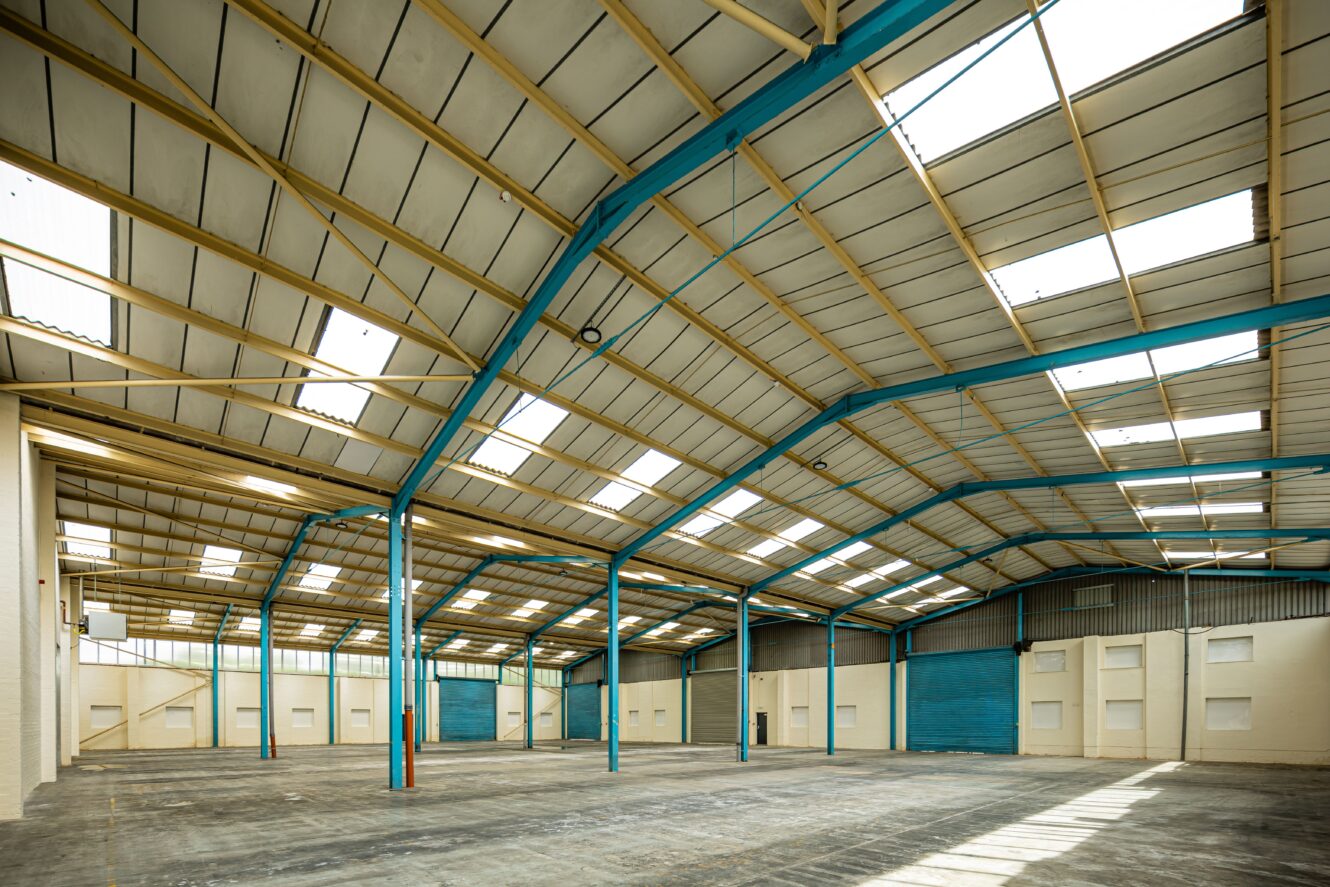 Large, empty warehouse interior with high ceilings, blue steel beams, exposed roof panels, and skylights allowing natural light to enter the open industrial space.