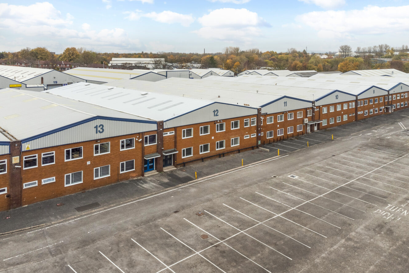 Rows of large warehouse buildings with numbered units and an empty parking lot in the foreground under a partly cloudy sky.