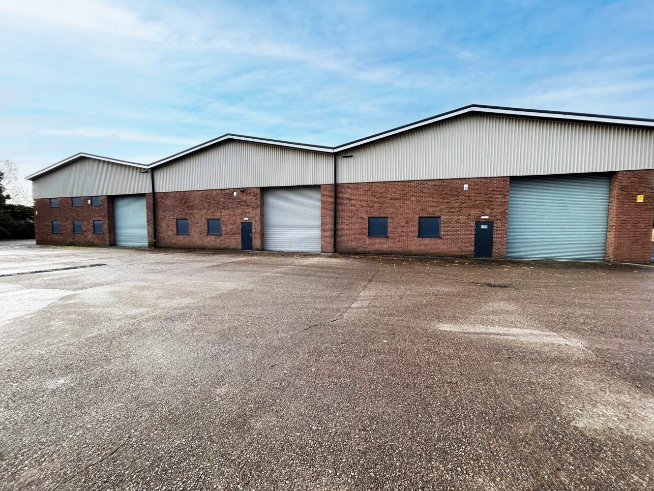 Large industrial warehouse building with three roller shutter doors and brick exterior, set on a paved lot under a blue sky.