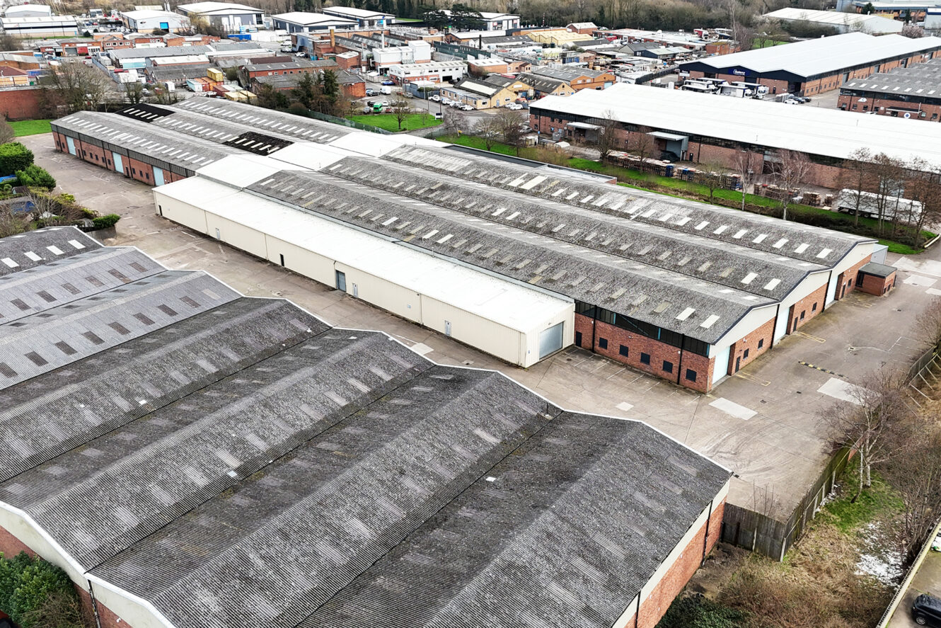 Aerial view of several large industrial warehouse buildings with gray roofs and brick walls, surrounded by concrete yards and adjacent factories.