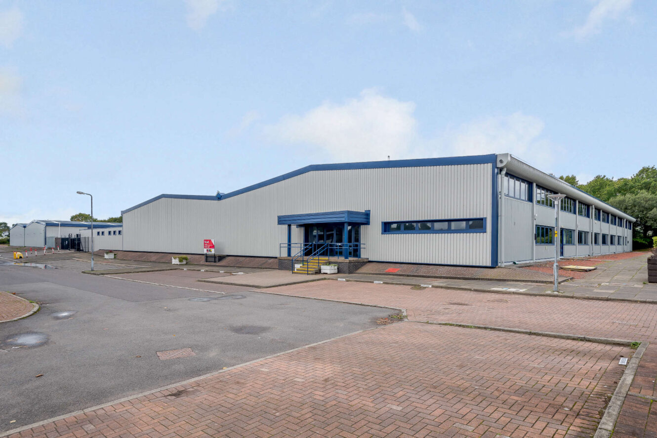 A large, modern industrial warehouse with blue and gray exterior, surrounded by an empty parking lot on a clear day.