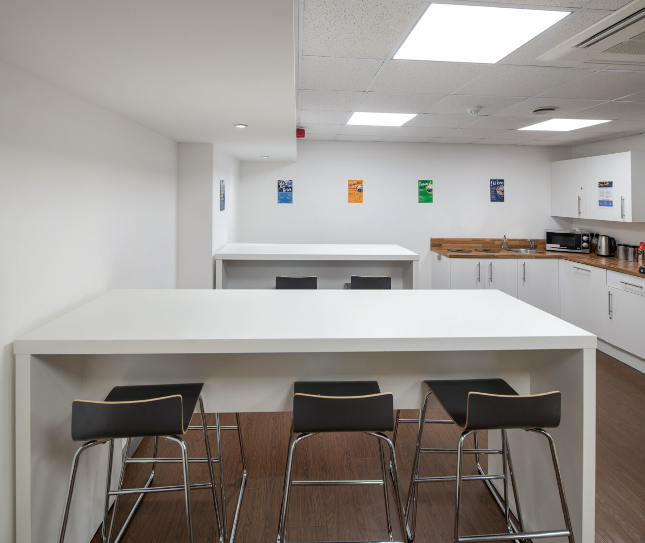 Modern office kitchen with white tables, black bar stools, white cabinets, countertop appliances, and colorful posters on the walls. The space is clean and well-lit.