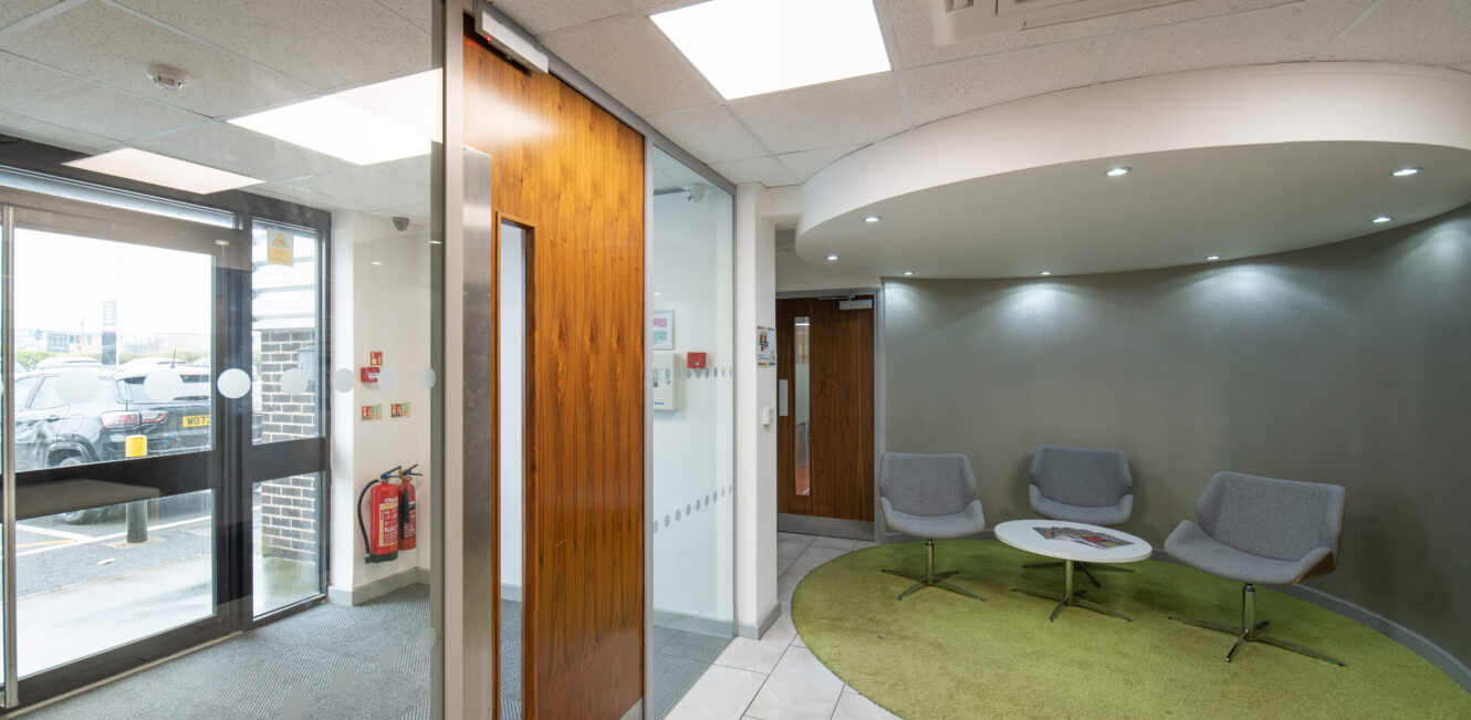 Modern office waiting area with four gray chairs around a white table on a green rug, next to a glass entrance door and fire extinguishers mounted on the wall.