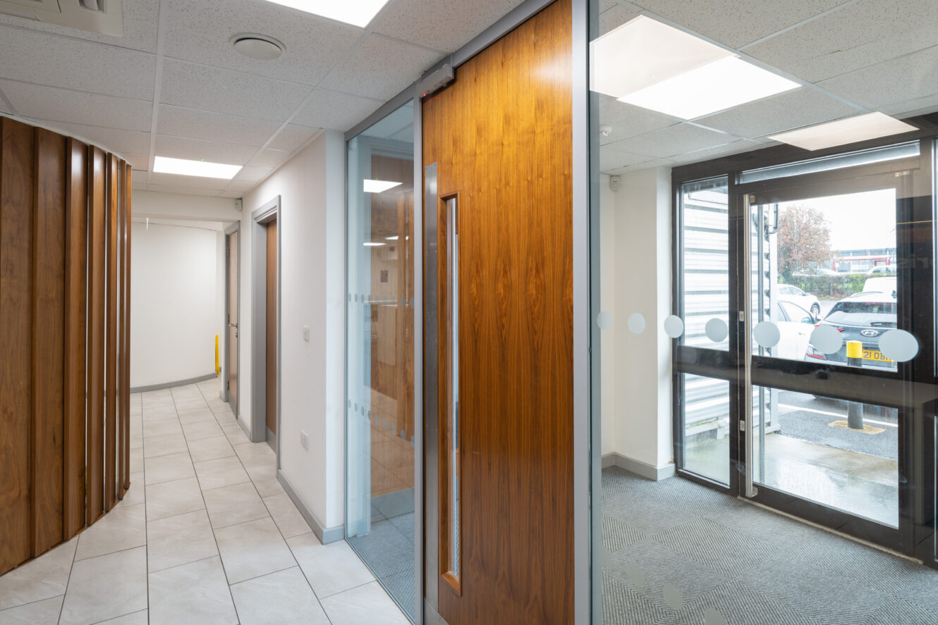 Office hallway with tiled floor, wooden doors, glass panels, and a glass entrance door showing parked cars outside.