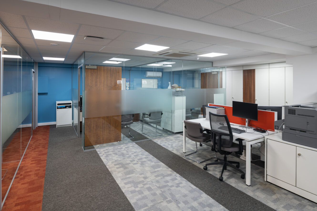 Modern office space with glass-walled rooms, desks with computers, office chairs, cabinets, and carpeted floors in different patterns.