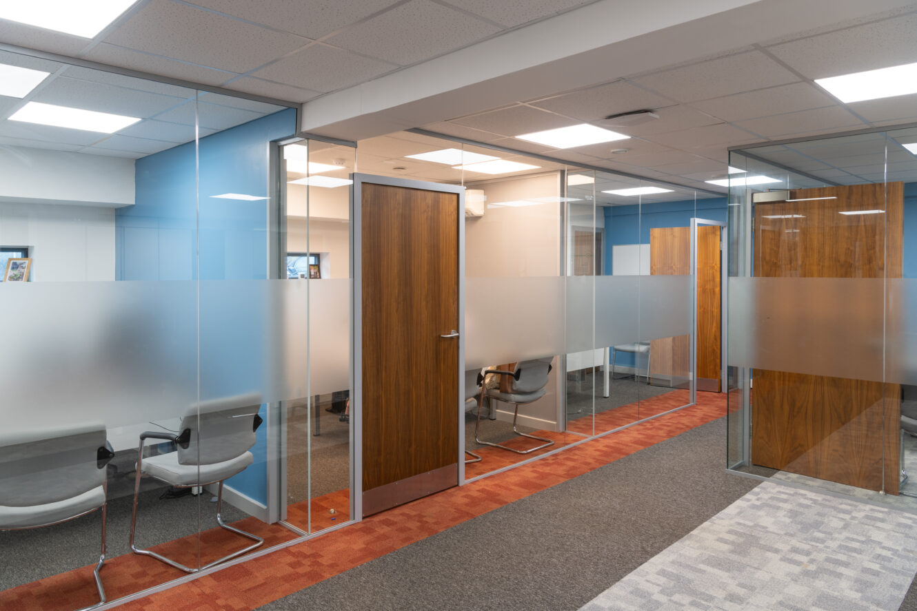 Modern office hallway with glass-walled meeting rooms, wooden doors, gray chairs, and a mix of carpet and tile flooring under fluorescent lighting.