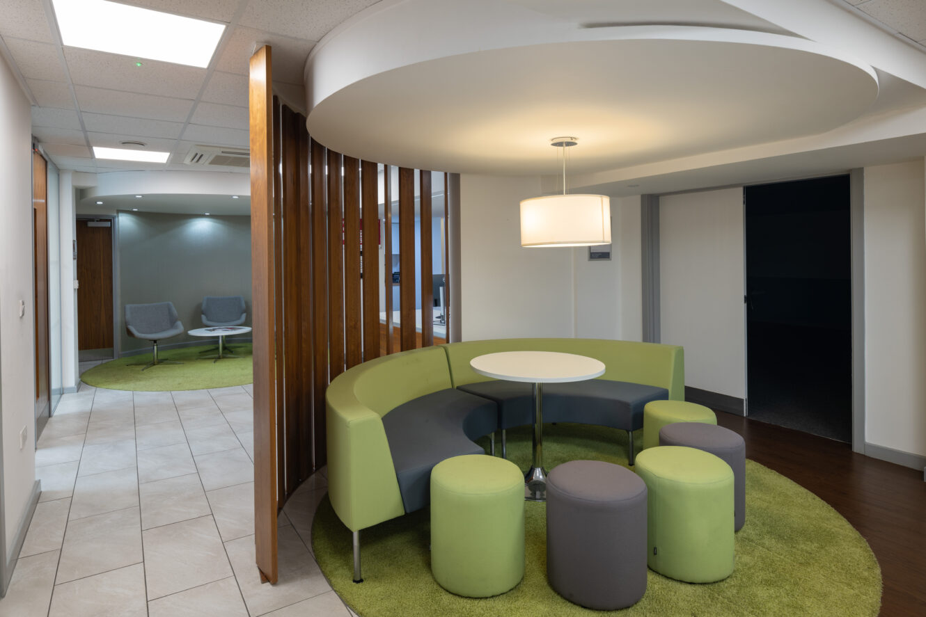 Modern office lounge with green and gray curved seating, round white table, cylindrical stools, and overhead light on a green rug, separated by vertical wooden slats.