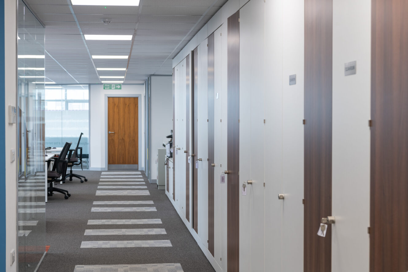 A modern office hallway with closed lockers on the right and a row of empty chairs at desks on the left, leading to a wooden door at the end.