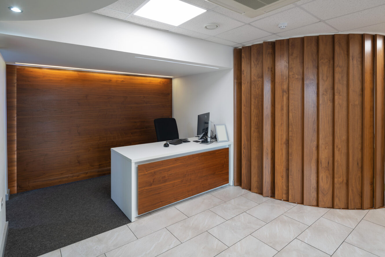 A modern reception desk with a computer and chair, set against a wooden accent wall and adjacent to a curved wooden partition, on a tiled floor.