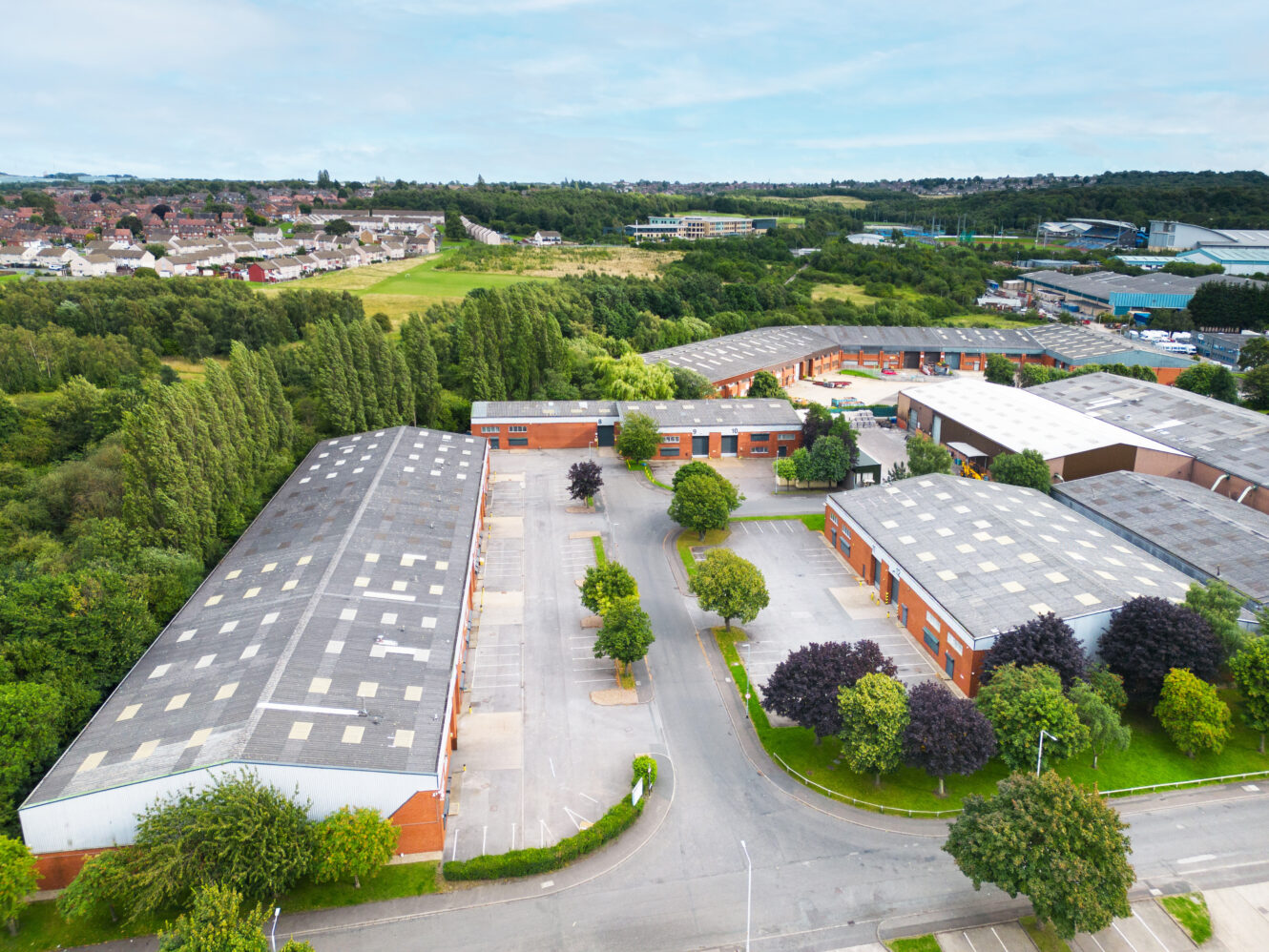 Aerial view of an industrial park with several large warehouse buildings, parking lots, and surrounding greenery under a partly cloudy sky.