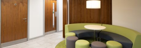 A waiting area with a curved green sofa, round white table, gray stools, overhead lamp, and wooden doors, including one with a restroom sign.
