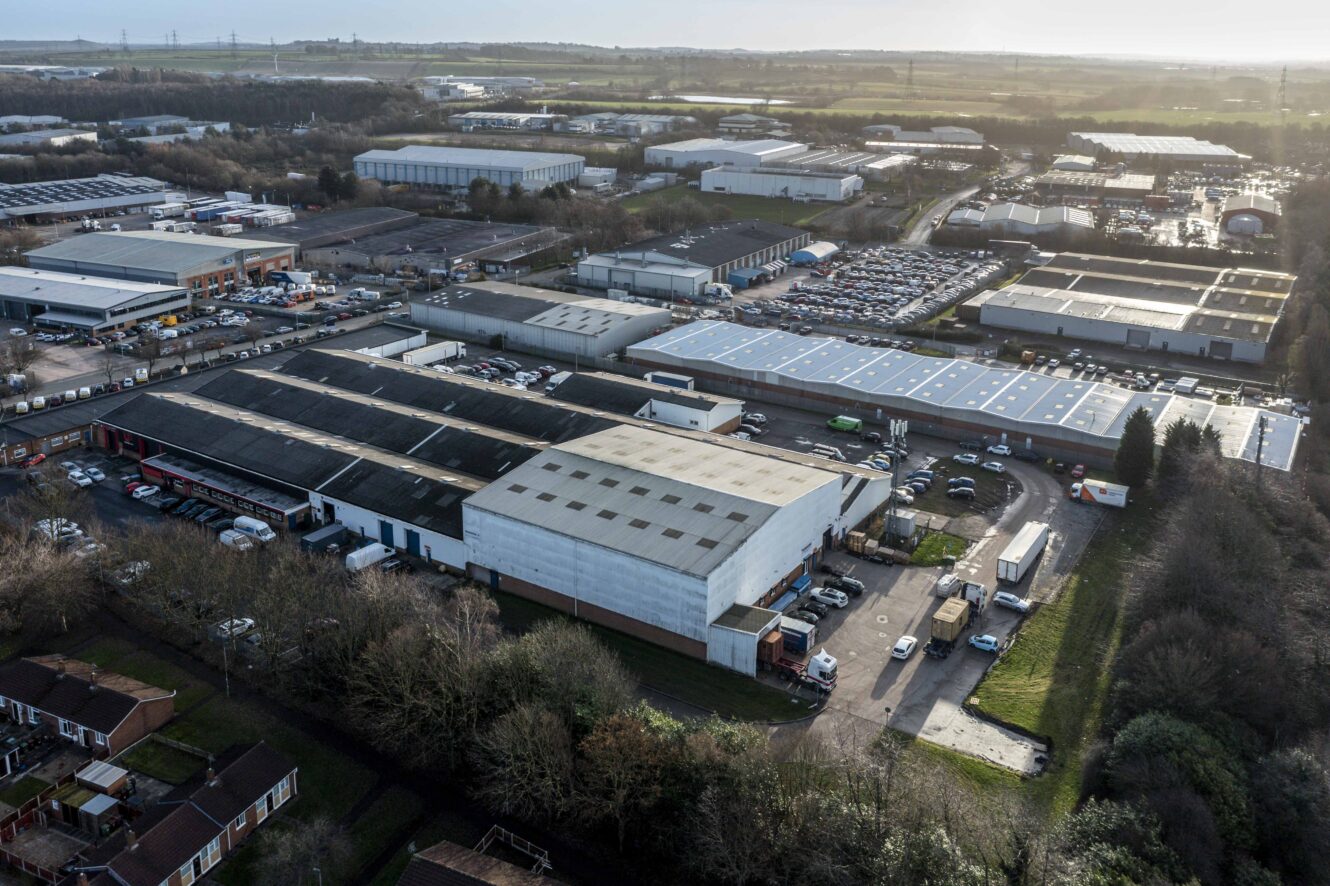 Aerial view of an industrial estate with large warehouses, parking lots, and surrounding roads bordered by trees and houses.