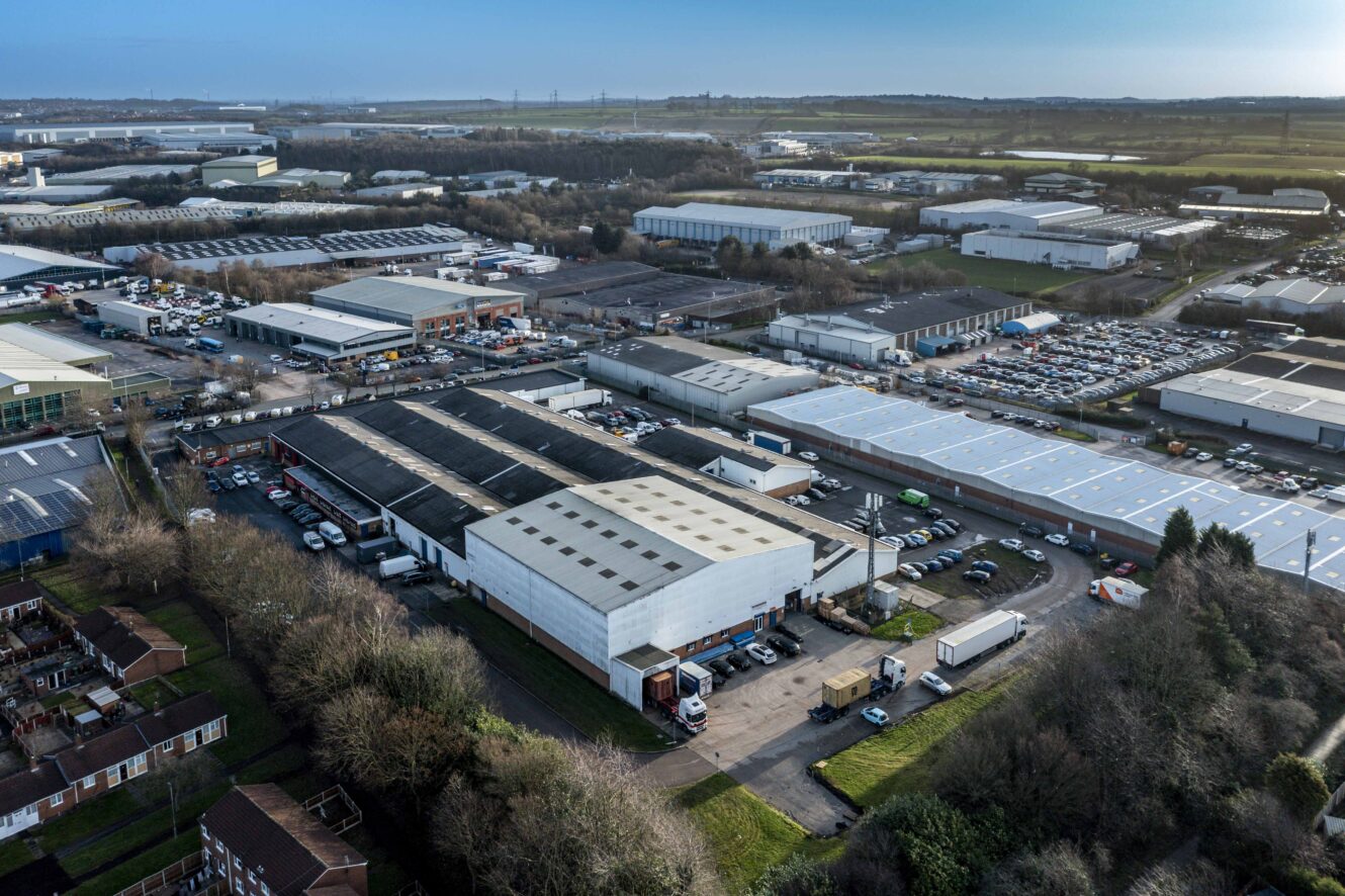 Aerial view of an industrial estate with multiple warehouses, parking lots, trucks, and nearby residential houses, surrounded by trees and open fields in the distance.