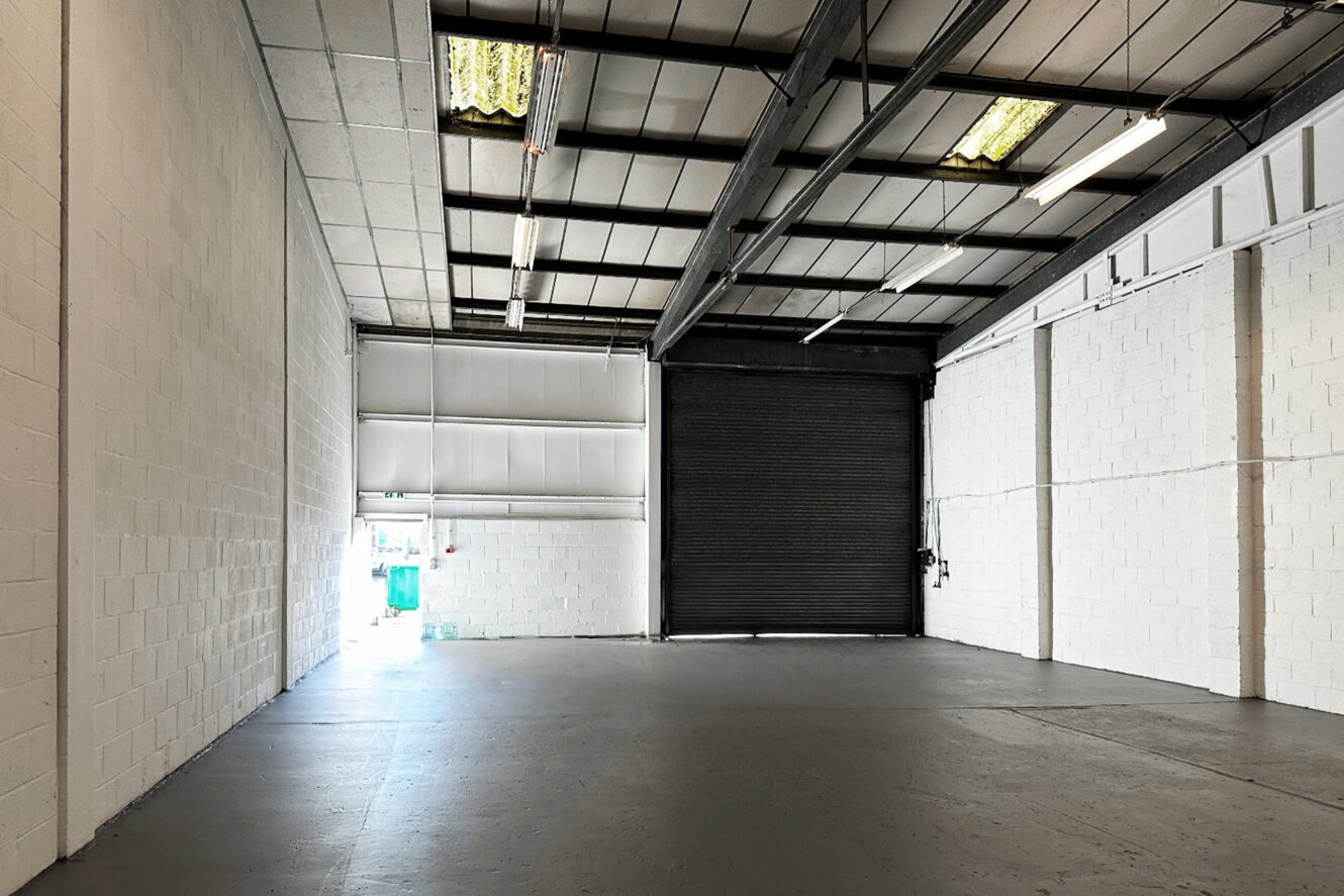 Empty industrial warehouse with white brick walls, high ceiling, exposed beams, fluorescent lights, and a large black roller shutter door partially raised revealing light from outside.