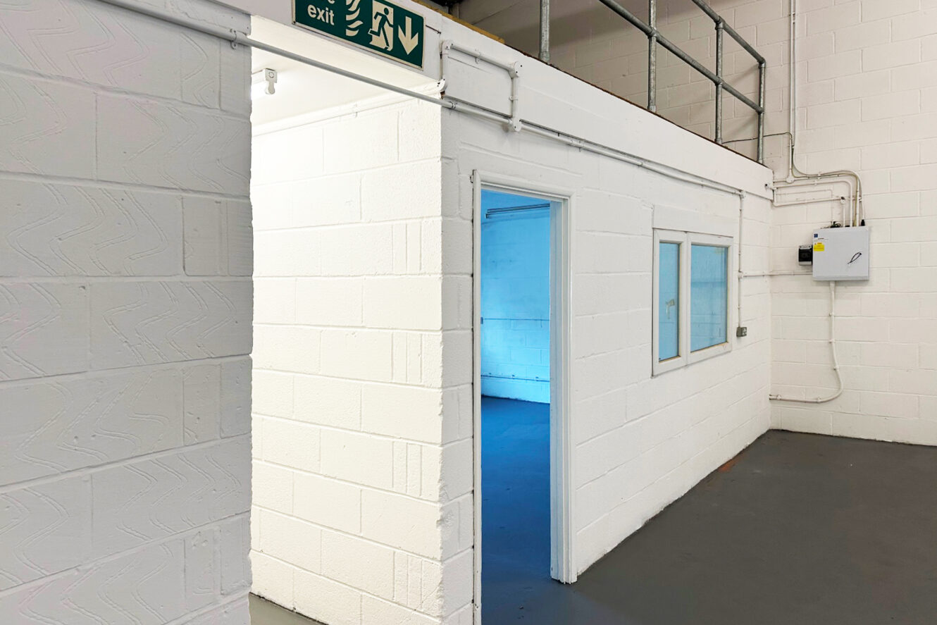 A white-walled room with an open doorway leads to another empty room. An exit sign is above the entrance, and there is a window and electrical box on the wall. The floor is gray.