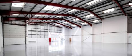Interior of a large, empty warehouse with clean, polished floors, white walls, red steel beams, and a partially open roller door letting in natural light.