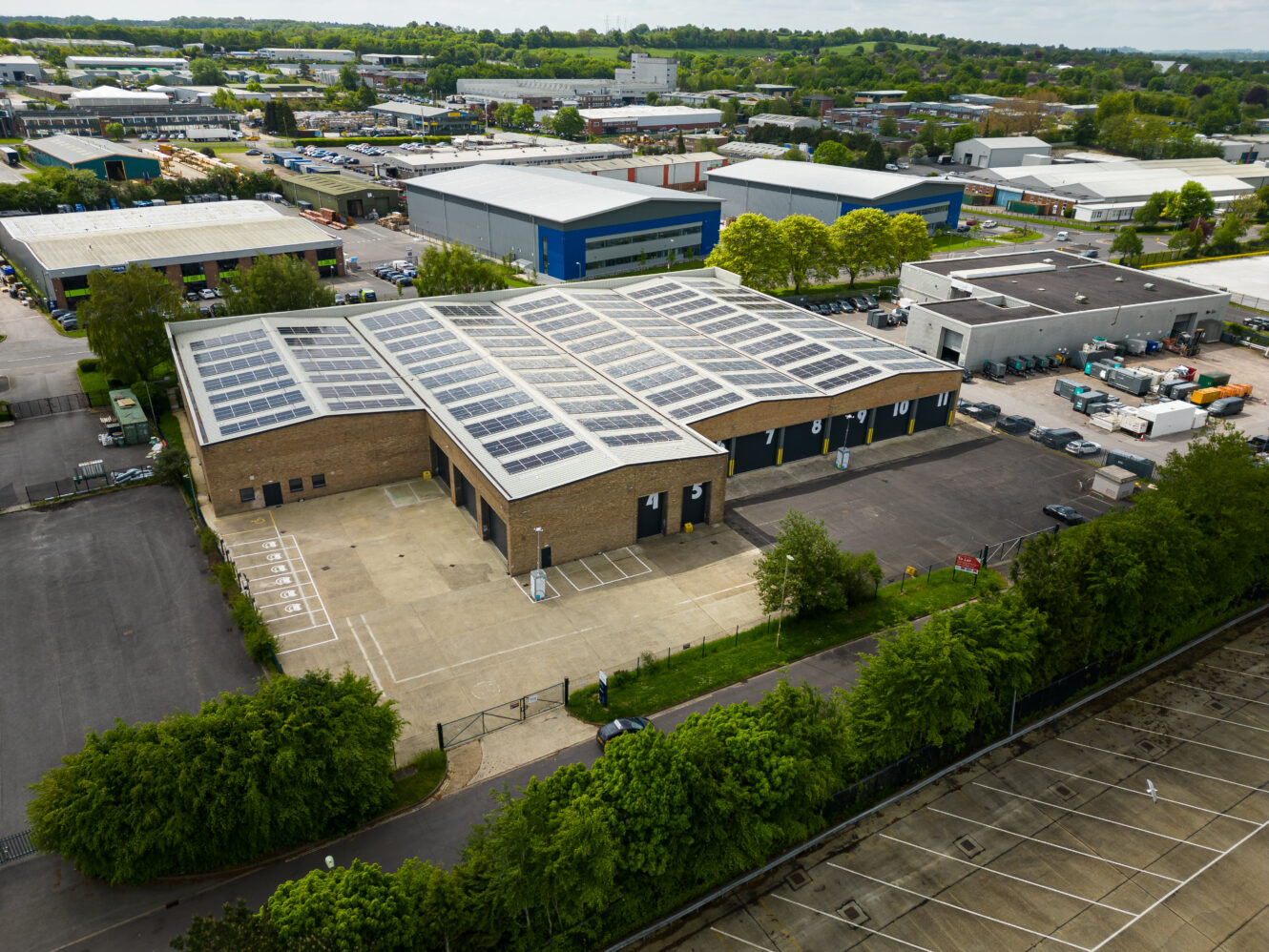 Aerial view of an industrial warehouse building with solar panels on the roof, surrounded by trees, parking spaces, and adjacent commercial buildings.