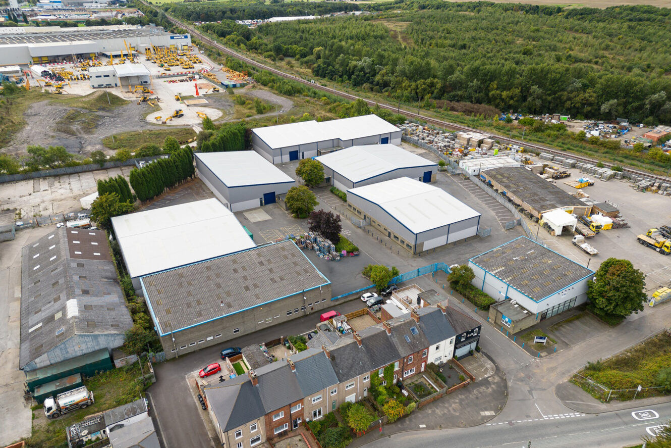 Aerial view of an industrial estate with warehouses, storage yards, and a row of residential houses alongside a main road, surrounded by greenery and a railway line.
