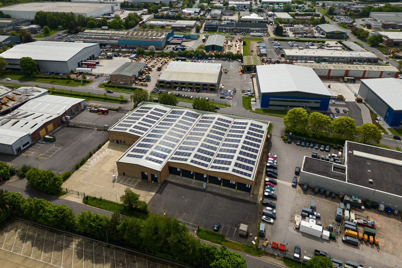 Aerial view of an industrial area featuring a large warehouse with solar panels on its roof, surrounded by parking lots, roads, and other commercial buildings.