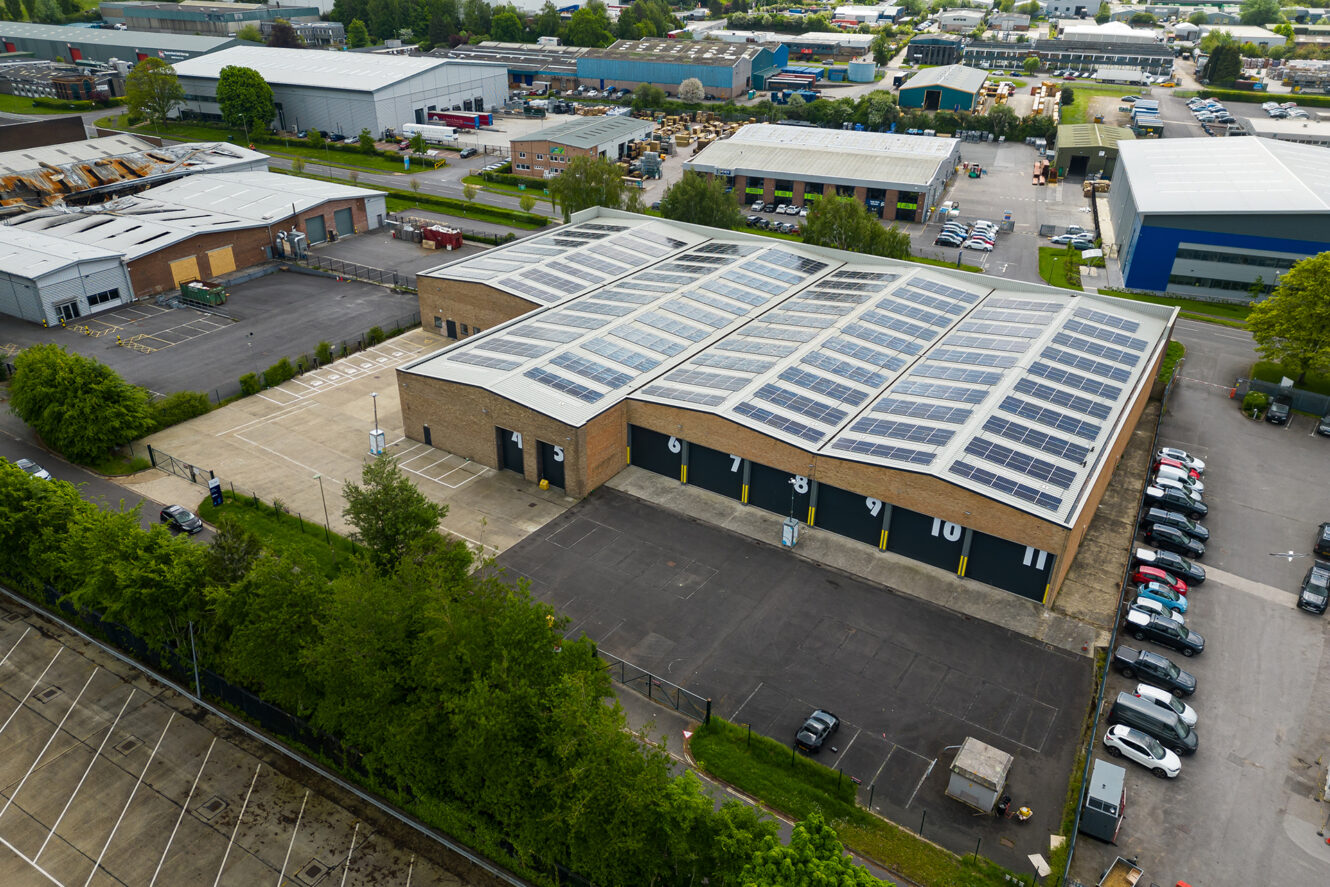 Aerial view of an industrial building with solar panels on the roof, surrounded by parking lots, trees, and nearby warehouses.
