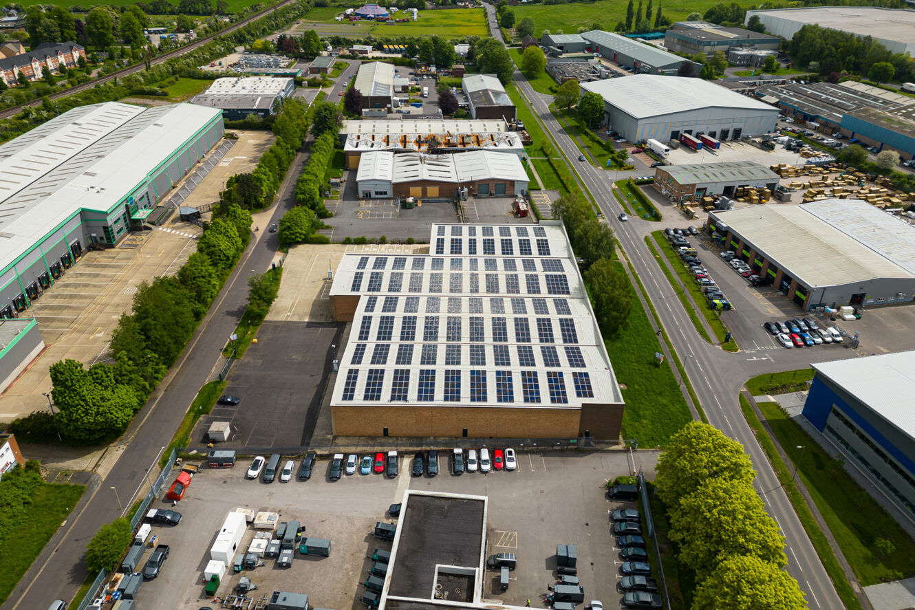 Aerial view of an industrial park with multiple warehouses, one building prominently featuring solar panels on its roof, surrounded by parking lots, roads, and green areas.
