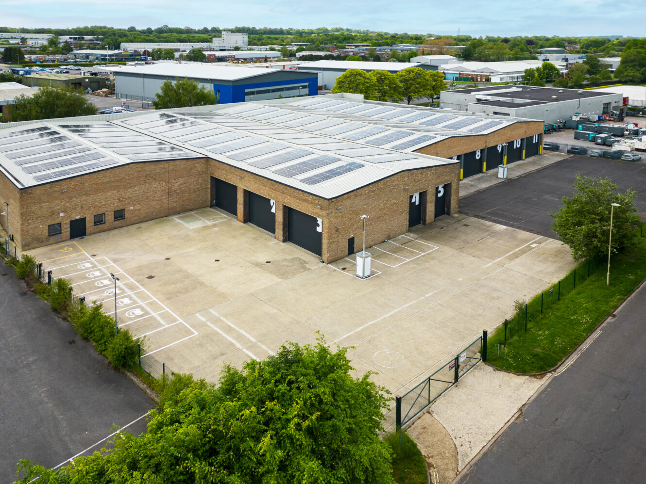 Aerial view of a modern industrial warehouse building with numbered loading bays, solar panels on the roof, and an adjacent parking area.