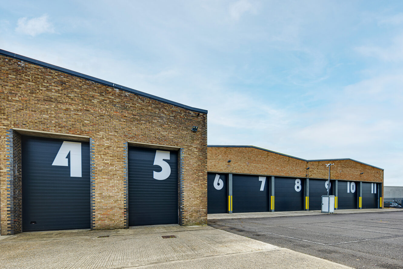 Row of brick industrial buildings with large black roller doors numbered 4 to 10, set against a clear sky and empty paved area in front.