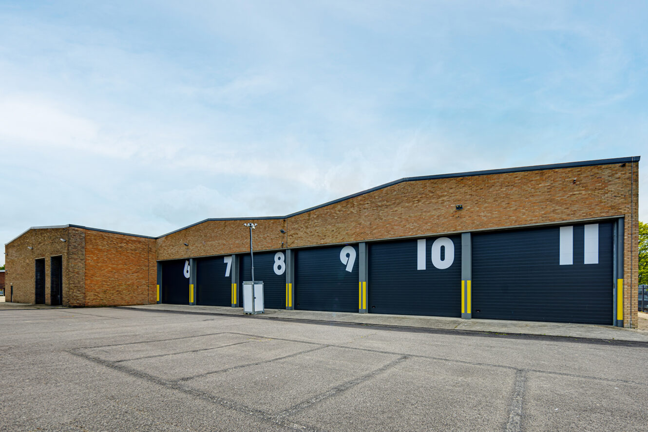 A row of brick industrial garages with black doors numbered 6 to 11 in large white digits, set in a paved lot under a clear sky.