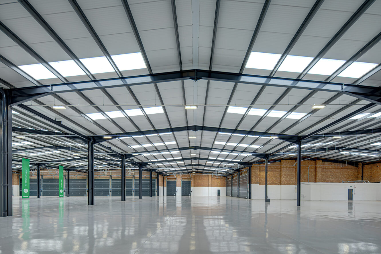 Large, empty industrial warehouse with polished concrete floors, exposed steel beams, brick walls, and skylights in the metal roof.