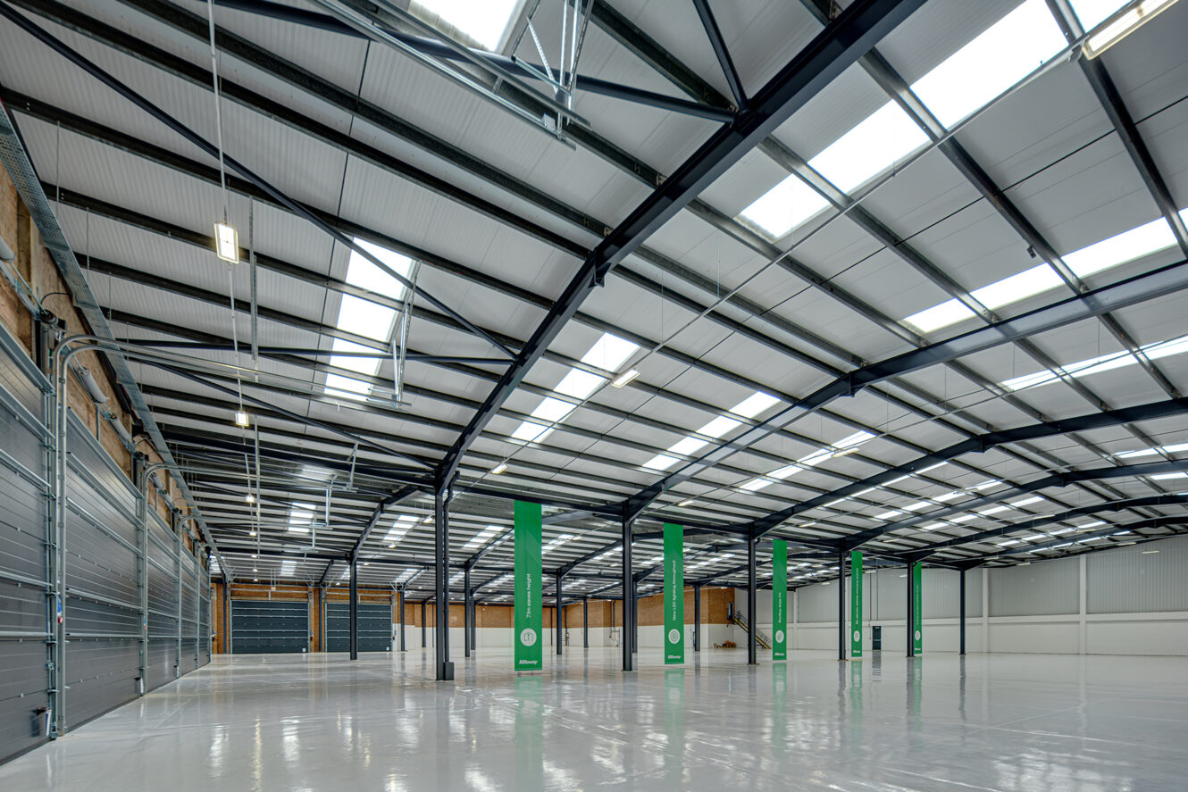 Wide-angle view of a large, empty warehouse with high ceilings, exposed steel beams, polished floors, and overhead skylights allowing natural light.