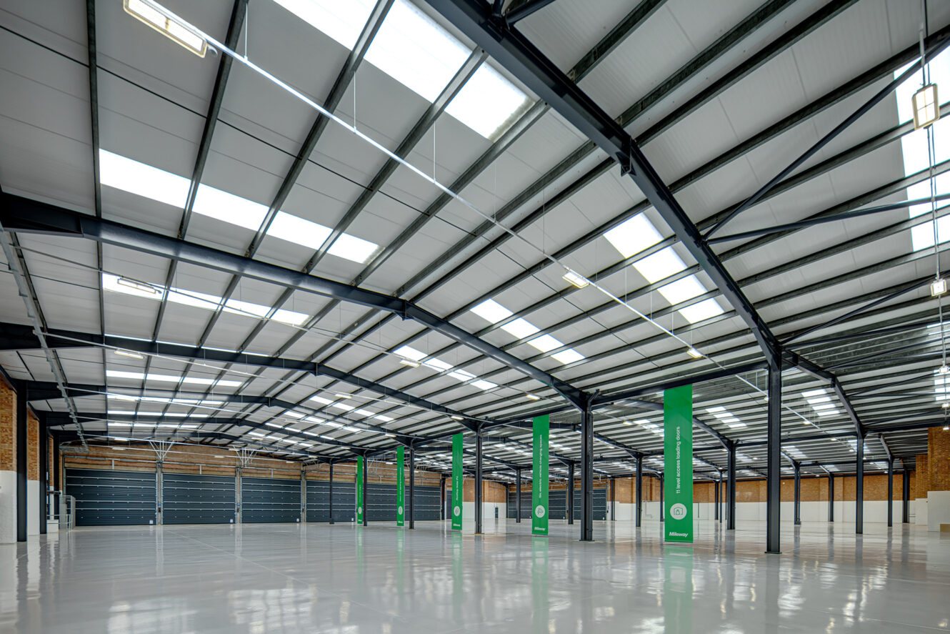 Large, empty industrial warehouse with polished floors, high ceilings, steel beams, brick walls, skylights, and green support columns.