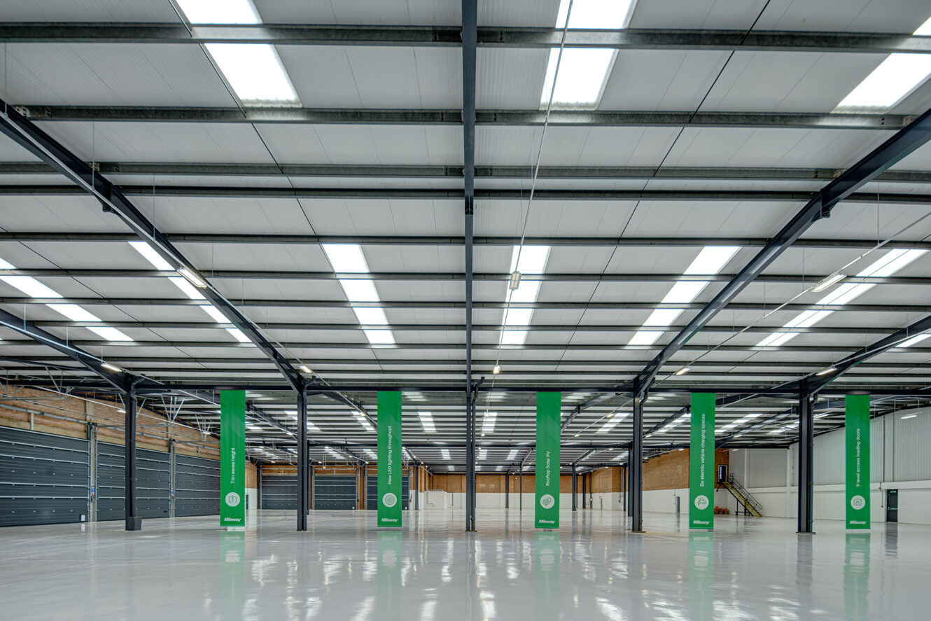 Interior of a large, empty warehouse with polished white floors, steel beams, green support columns, and bright overhead lighting.