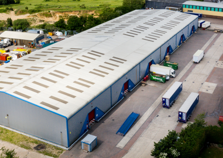 A large industrial warehouse with several loading bays, trucks, and trailers parked outside, surrounded by greenery and other buildings.