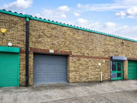 A brick industrial building with a closed metal roller shutter, a green metal door, and a green garage door under a partly cloudy sky.