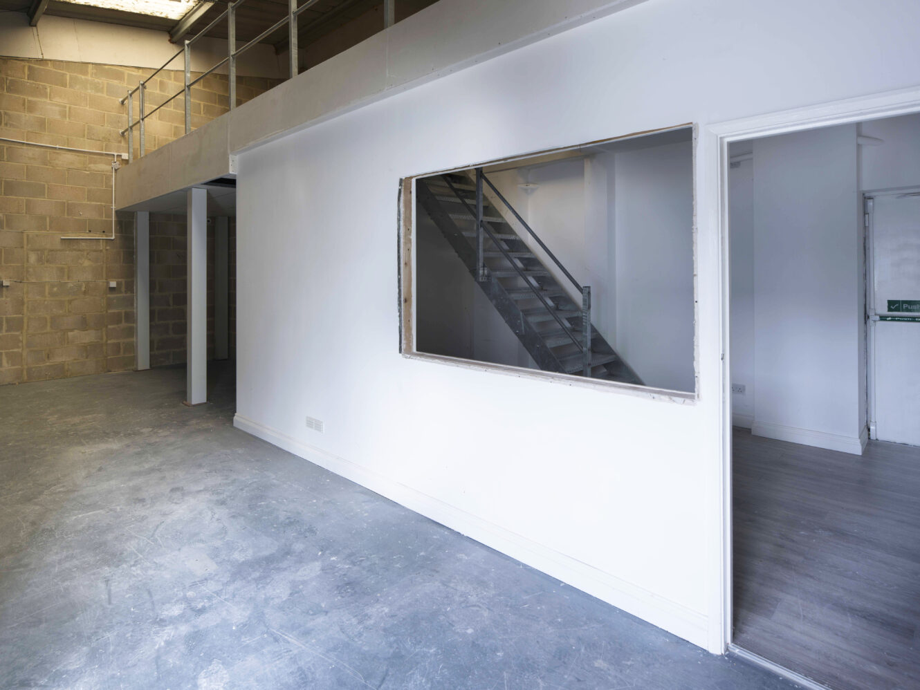 Empty industrial interior with concrete floor, white walls, a large window opening, and a staircase visible through an interior window and doorway.
