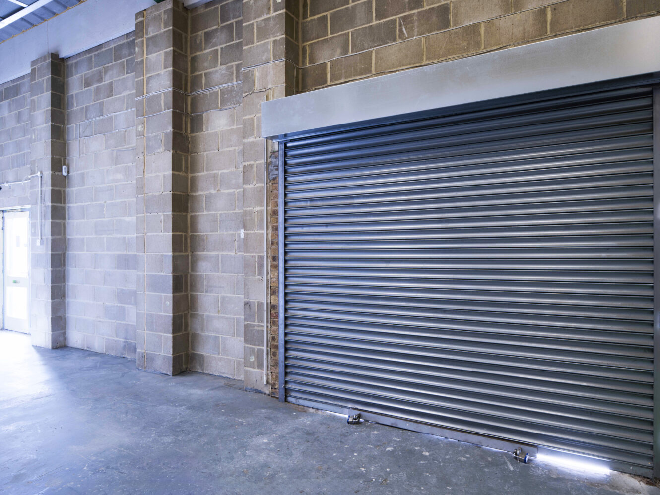 A closed metal roll-up garage door in an industrial building with concrete block walls and a concrete floor.