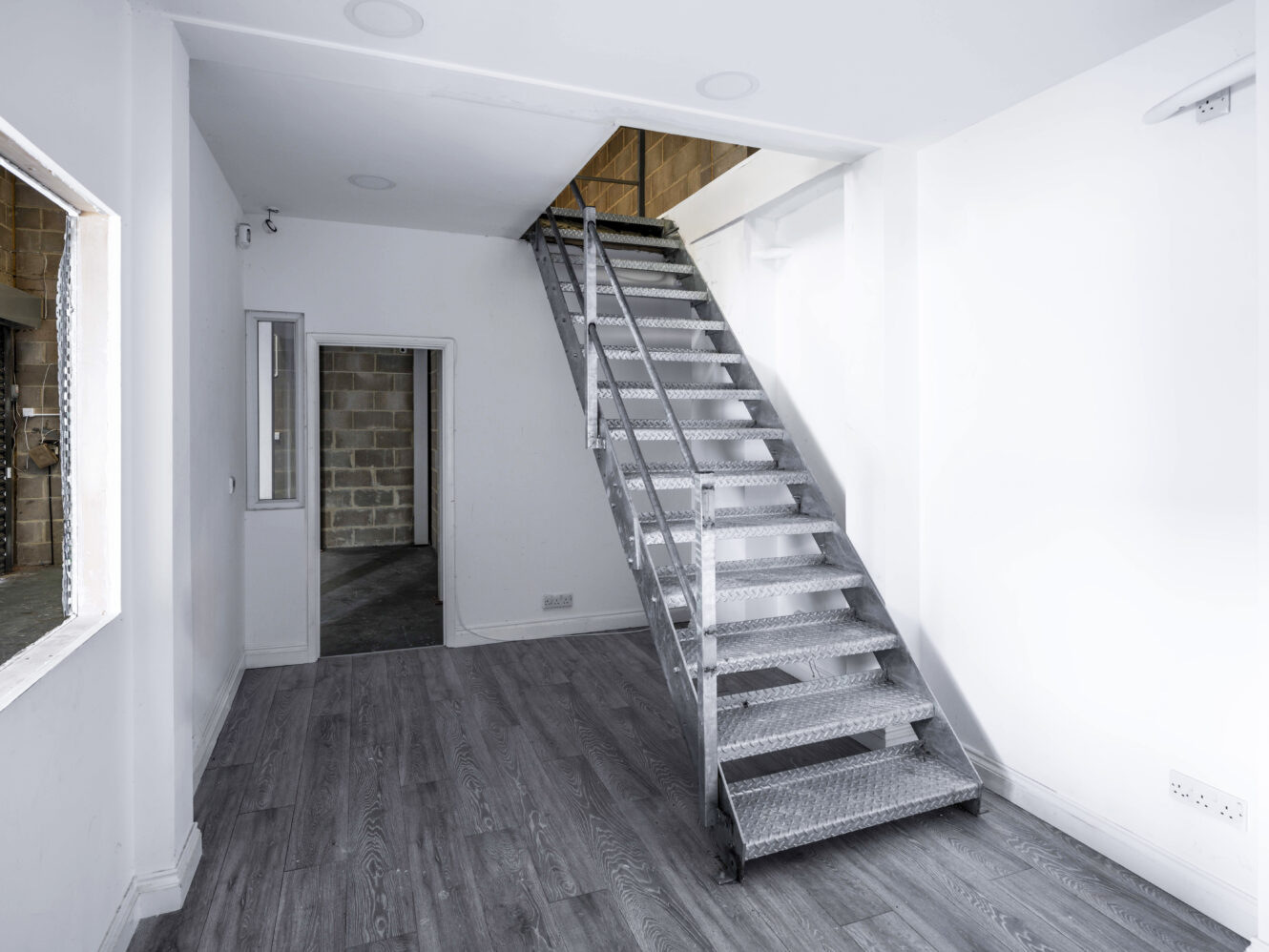 A metal staircase leads to an upper floor in a minimally furnished room with white walls and grey wood flooring. An open doorway and a window are visible in the background.