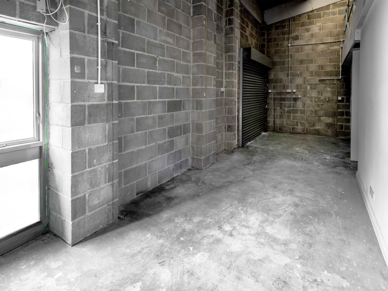 An empty industrial space with concrete block walls, a large window, exposed wiring, and a rolled-down metal shutter door.