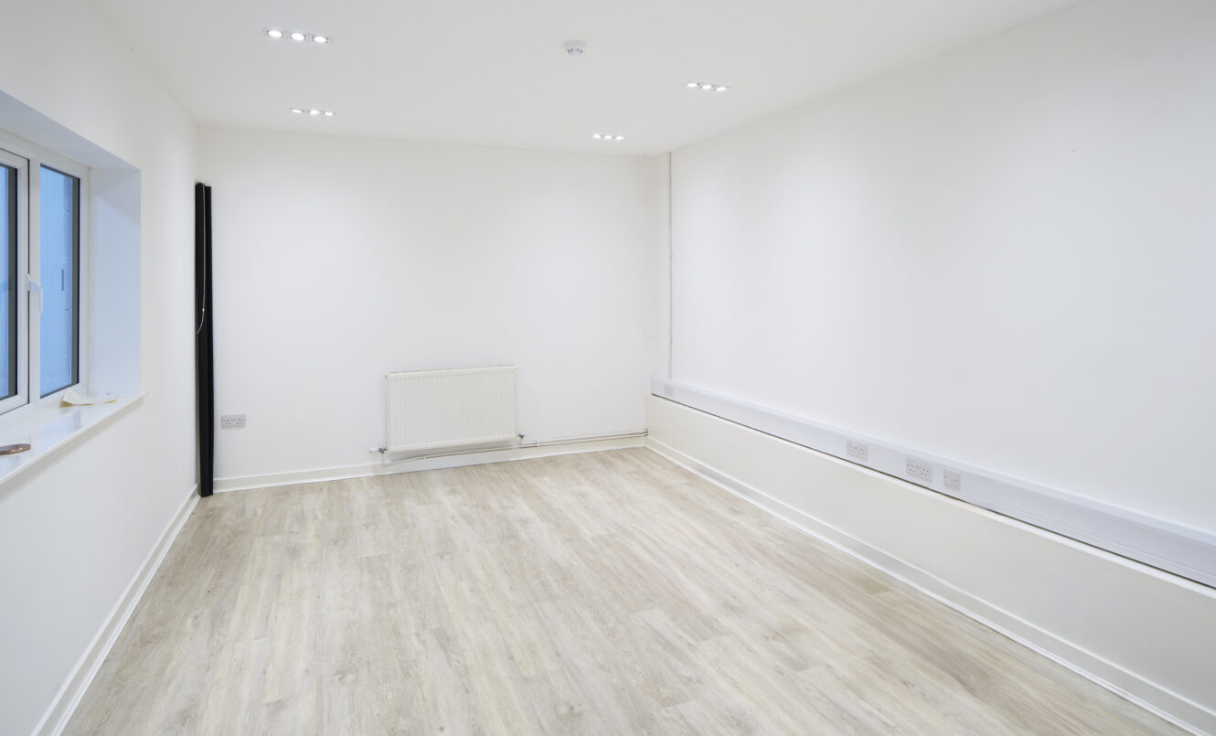Empty, unfurnished room with white walls, light wood flooring, a radiator, electrical outlets, and a window on the left wall. Ceiling lights are turned on.