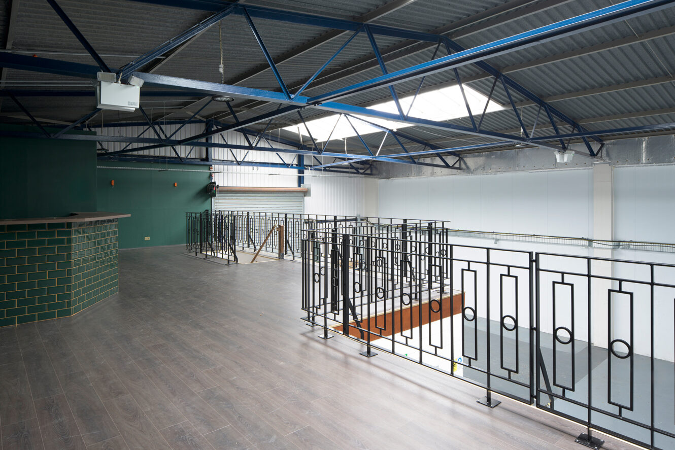 Interior of a modern, industrial loft with metal railings, exposed beams, green tiled bar, and dark wood flooring, overlooking an open space below.