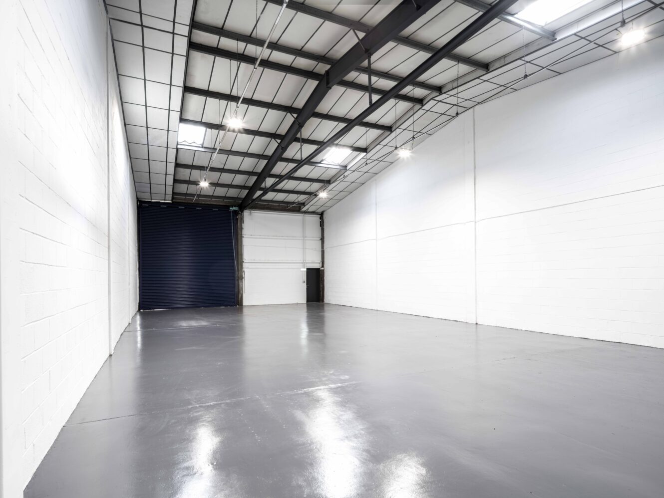 Empty industrial warehouse interior with white walls, high ceiling with exposed beams, polished concrete floor, and a large closed roller door at the back.