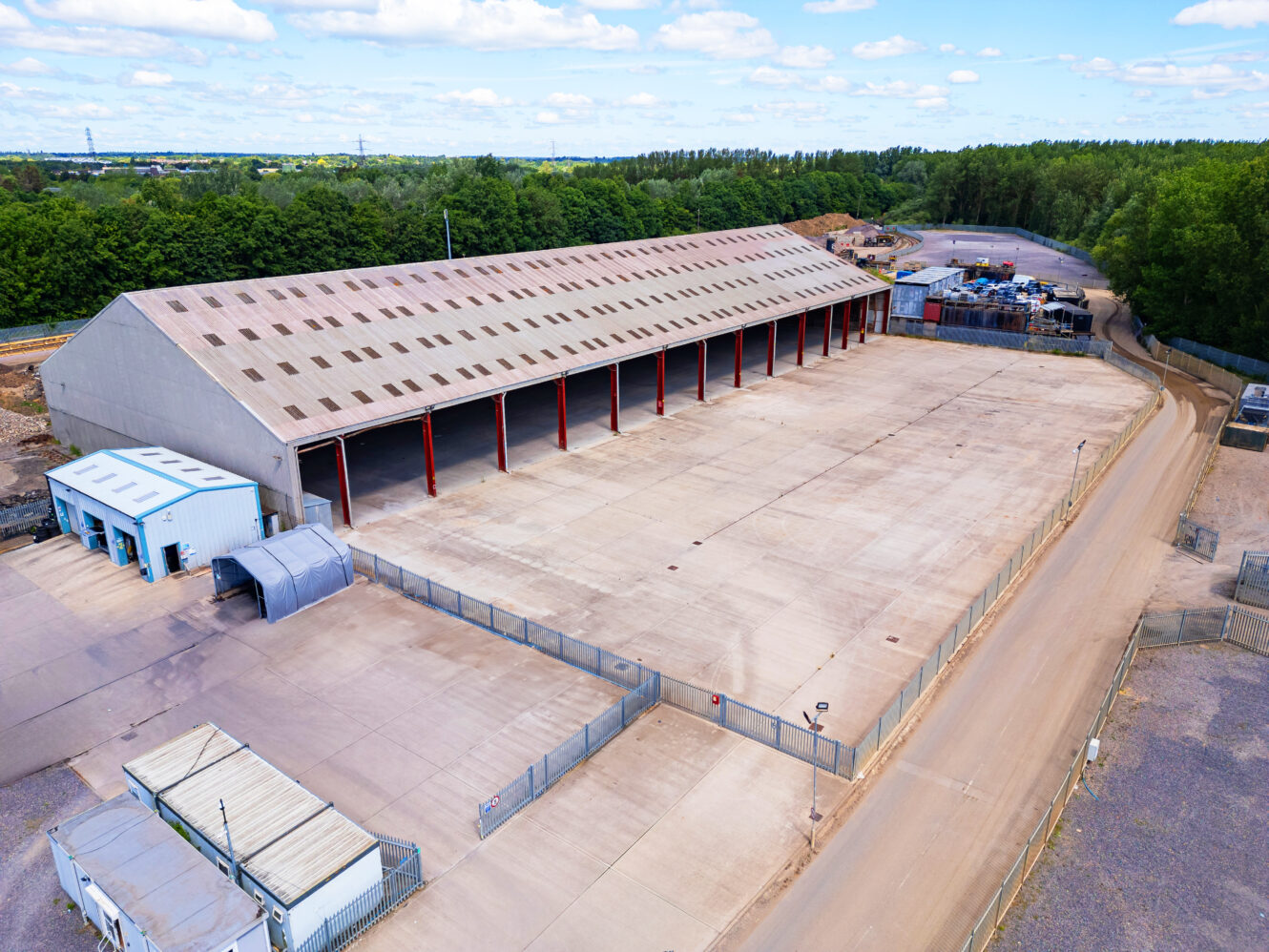 A large, empty industrial warehouse with an open front is surrounded by a fenced lot and adjacent structures, set near a wooded area under a partly cloudy sky.