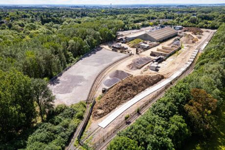 Aerial view of a construction site with large cleared areas, building materials, and surrounding trees and railway tracks.