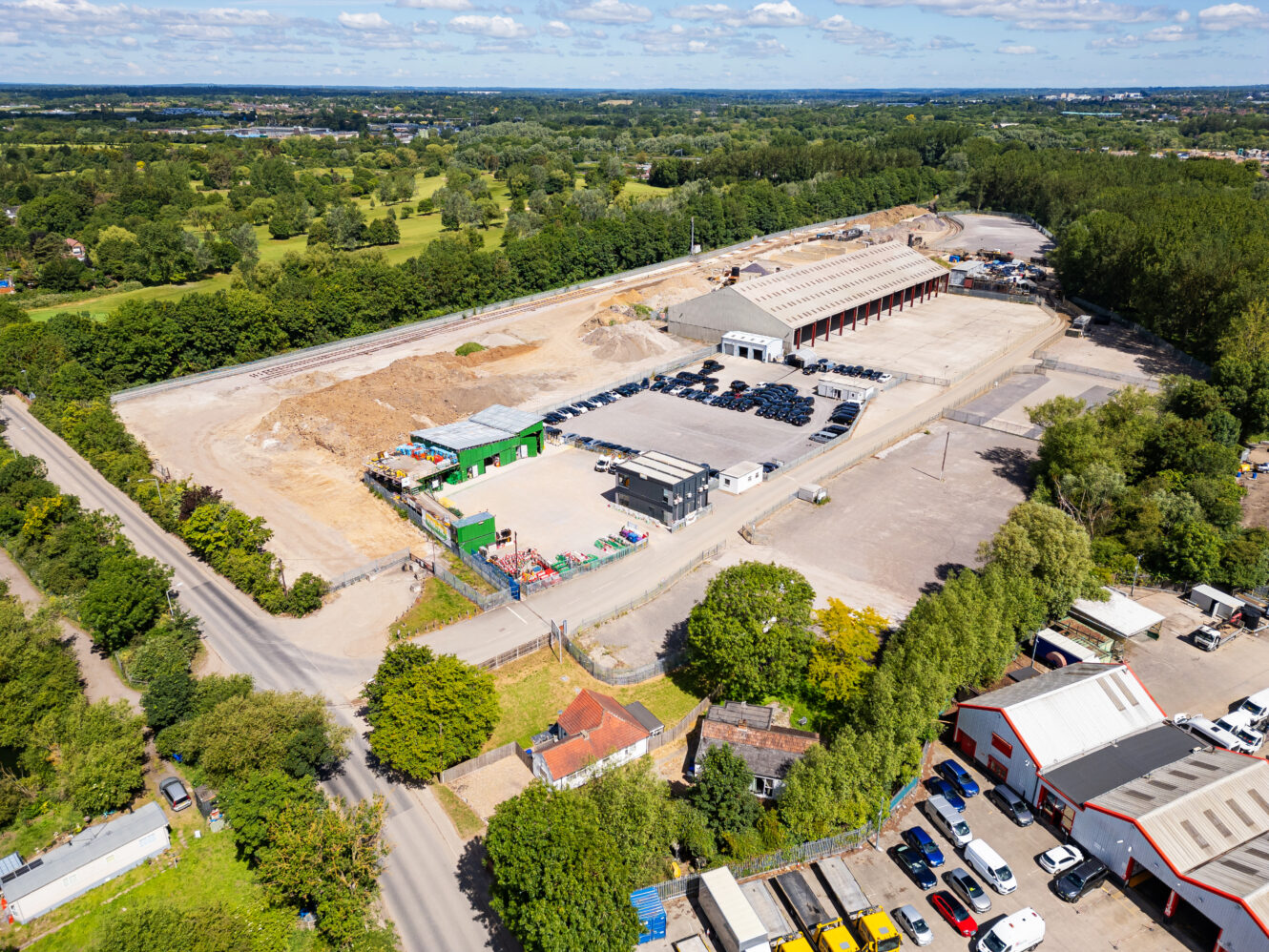 Aerial view of an industrial site with multiple warehouses, parked vehicles, and surrounding greenery under a blue sky with scattered clouds.