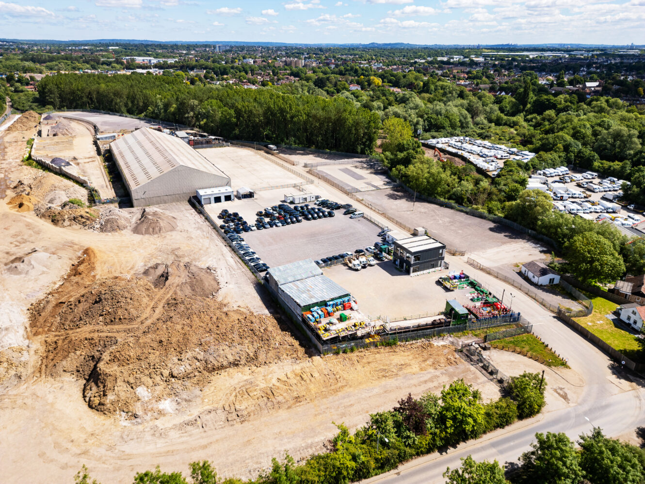 Aerial view of an industrial facility with parked vehicles, buildings, and surrounding construction areas, bordered by trees and adjacent to a residential area with trailers.
