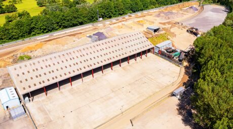 Aerial view of a large, empty warehouse with a sloped roof next to a construction or storage yard, surrounded by trees and open land.