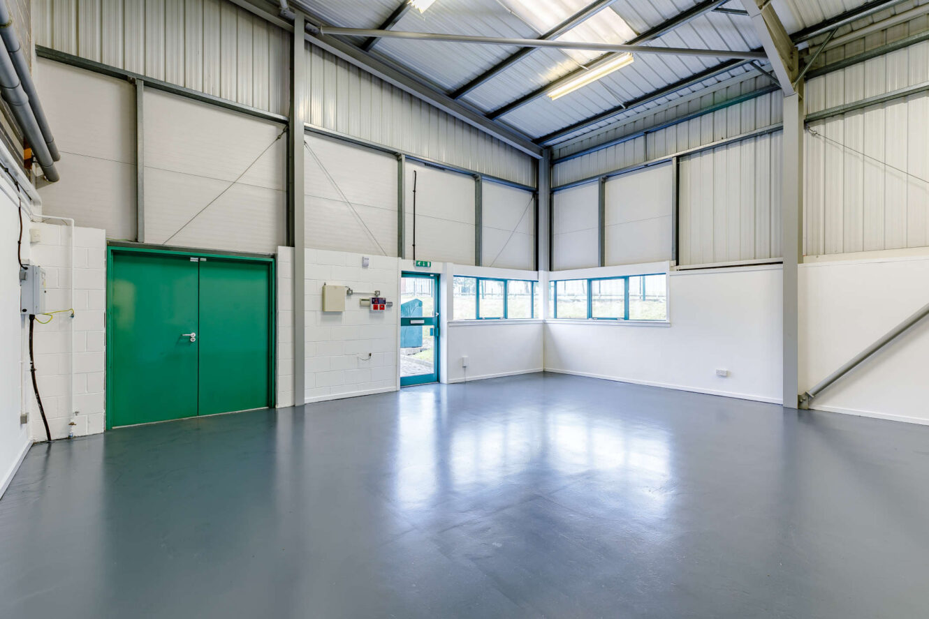 Empty industrial unit with grey floor, white and metal walls, green double doors, large windows, and exposed metal ceiling beams. Fluorescent lights are on.