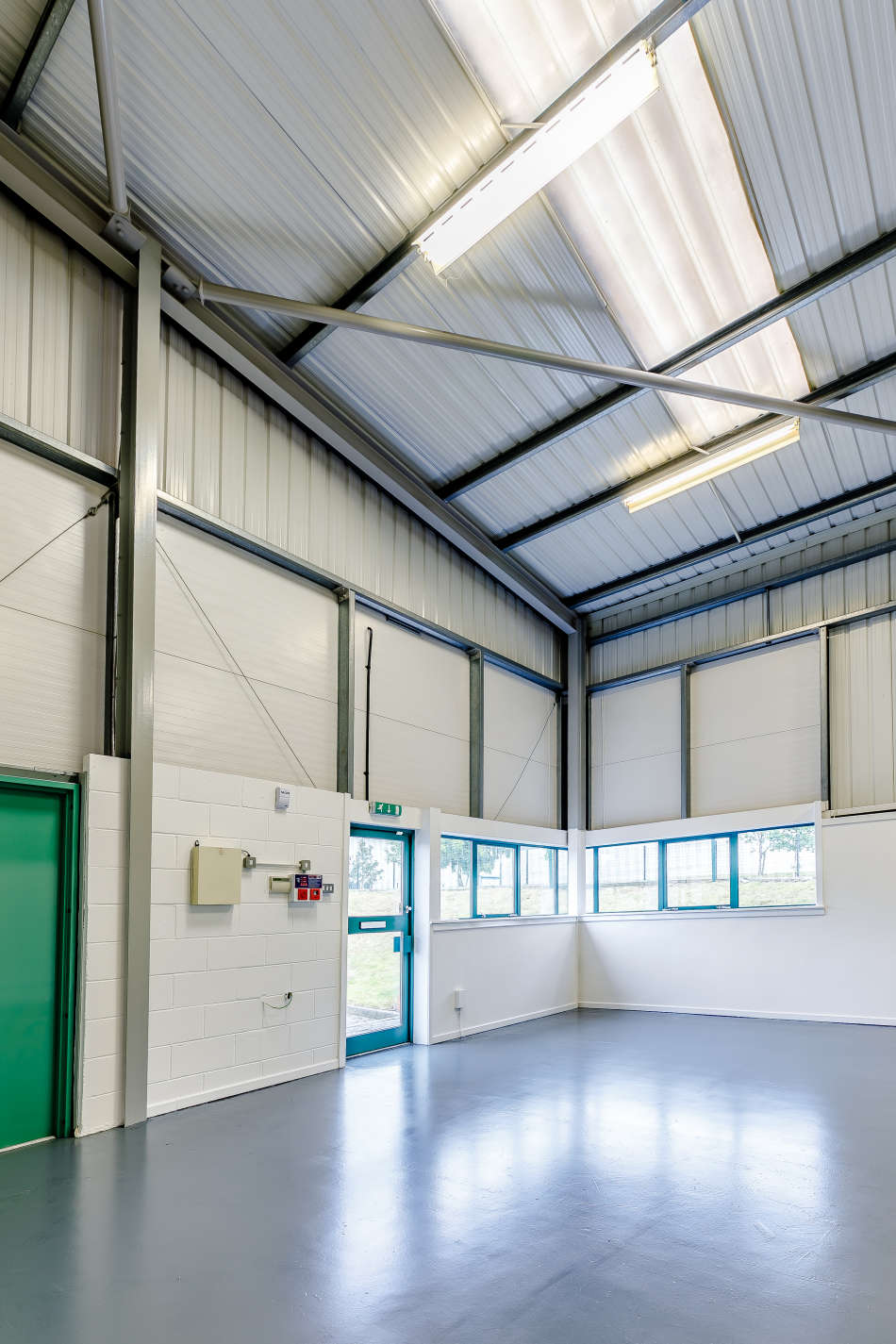 Interior of an empty industrial unit with high ceilings, metal beams, white walls, green doors, windows, and grey flooring. Fluorescent lights are on the ceiling.