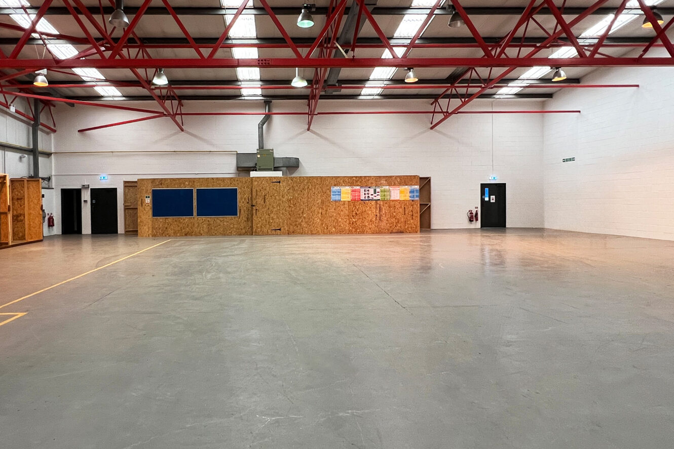 Large empty warehouse with concrete floor, exposed red ceiling beams, and plywood office structures along the back wall, under fluorescent lighting.