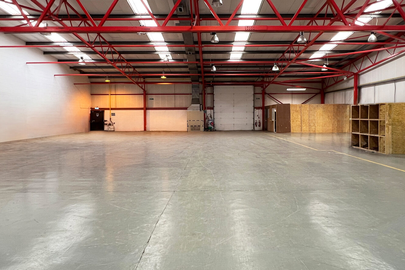 Spacious, empty warehouse with red ceiling beams, concrete floor, and some wooden storage units along the right wall. Overhead lights are on.